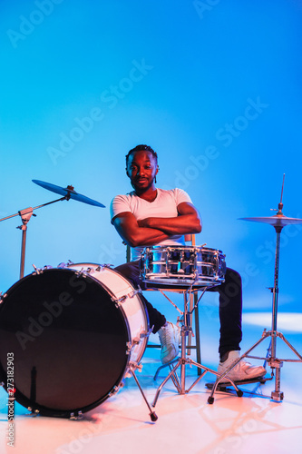 Young african-american jazz musician or drummer playing drums on blue studio background in trendy neon lights. Concept of music, hobby, inspirness. Colorful portrait of joyful attractive artist. photo