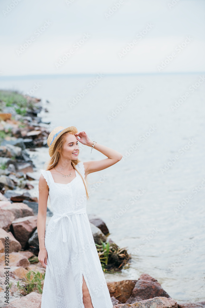 cheerful young blonde woman touching straw hat and looking at sea