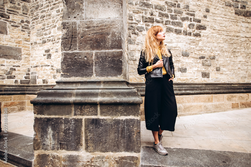 Young blonde with a backpack walking along a narrow cobbled street of Prague, Czech Republic. Stylish blonde girl in a leather jacket.