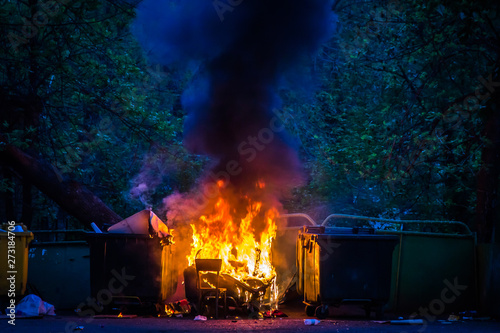 Burning trash container and old wood chair with other garbage. Plastic garbage can has almost fused