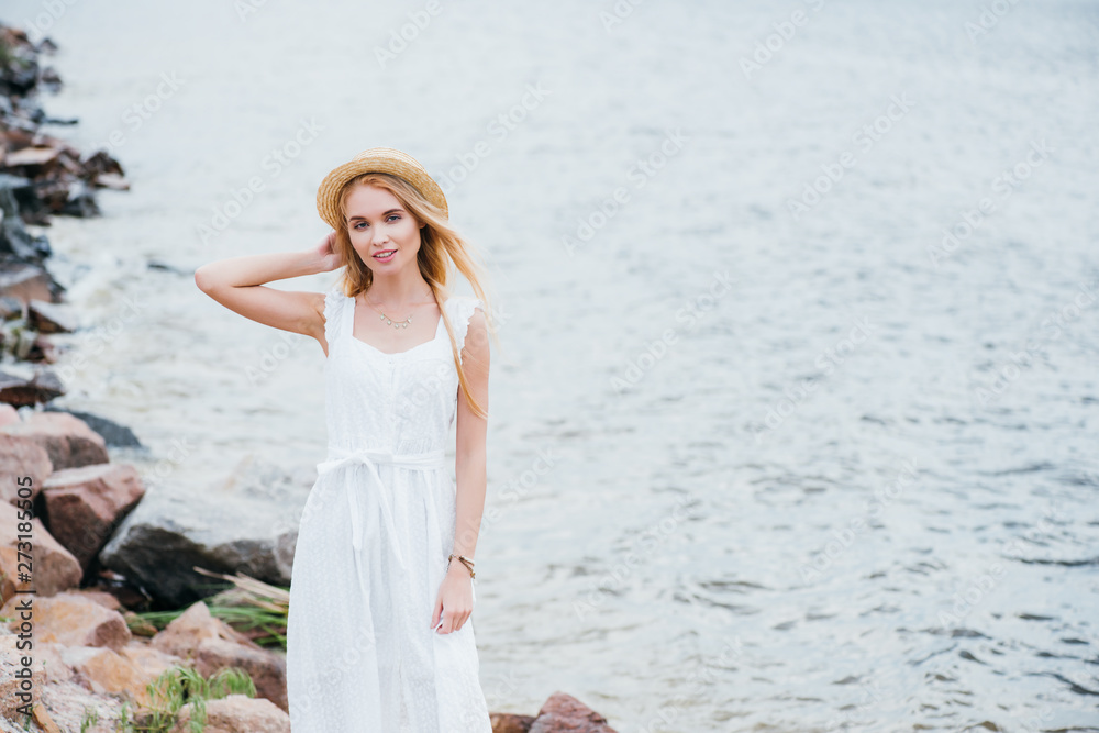 cheerful young blonde woman touching straw hat and looking at camera near sea cheerful young blonde woman touching straw hat and looking at camera near sea