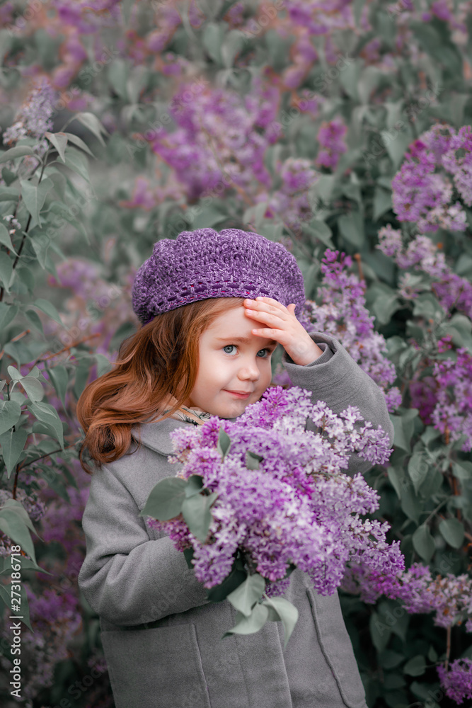 Little red-haired girl stands in lilac bushes with a bouquet