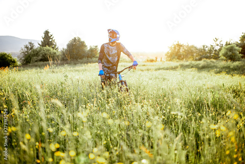 Professional well-equipped cyclist walking with bicycle on the beautiful green field on the mountains during the sunset