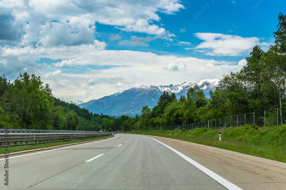 Autobahn or highway in the mountains with clear marking surrounded by vibrant green trees under blue sky. Stunning view and a snow-covered mountain in the background. The Alps, Austria Autobahn or highway in the mountains with clear marking surrounded by vibrant green trees under blue sky. Stunning view and a snow-covered mountain in the background. The Alps, Austria
