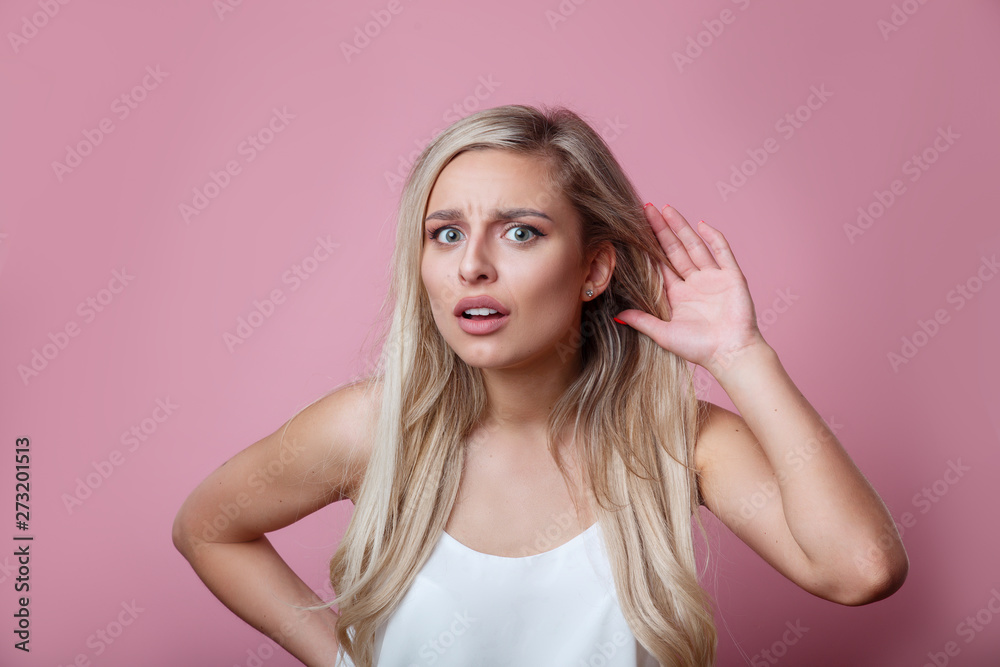 Beautiful young woman puts a hand to the ear to hear better on a pink background