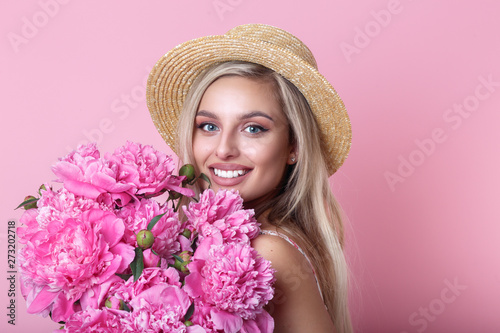Close-up portrait of beautiful young woman in straw hat holding peonies bouquet over pink background