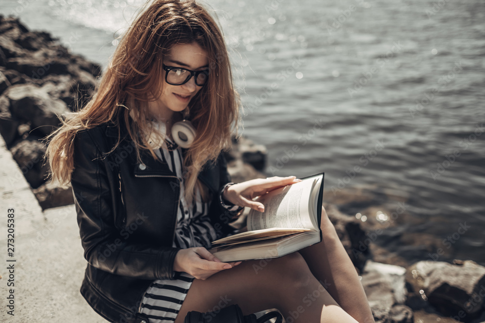 Outdoor Summer Portrait of Young Beautiful Girl in Black Leather Jacket and Glasses Reads the Book Near the Lake