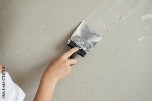 cute girl holds a putty knife over a gray wall.
