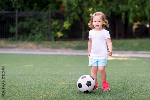 Blonde toddler girl in blue shorts and pink sneakers playing with soccer ball at football field or stadium outdoors. Green background. Child dreams, girl power and active childhood concept. Copy space