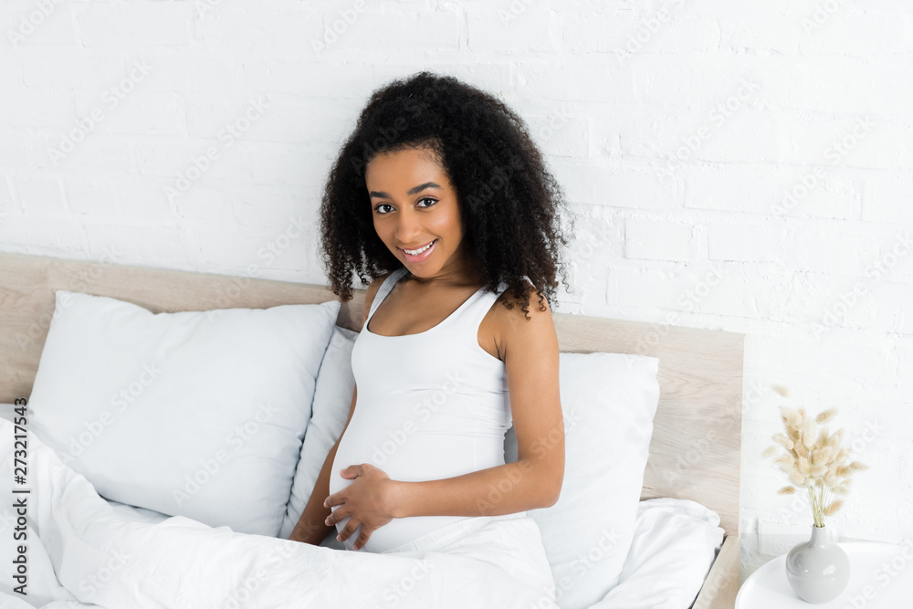pregnant african american woman looking at camera, sitting on bed in room pregnant african american woman looking at camera, sitting on bed in room