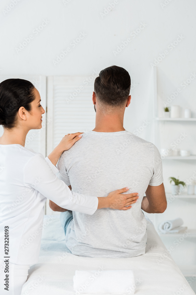back view of man sitting on massage table near attractive healer back view of man sitting on massage table near attractive healer