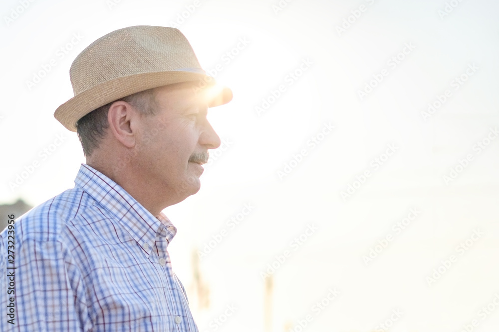 Retired senior hispanic man with hat standing and smiling at sunny day. Profile view