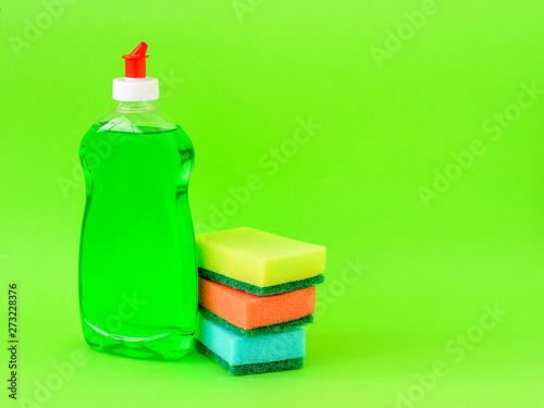 фотографія Transparent bottle with green dishwashing liquid and three foam sponges of different colours on a green background