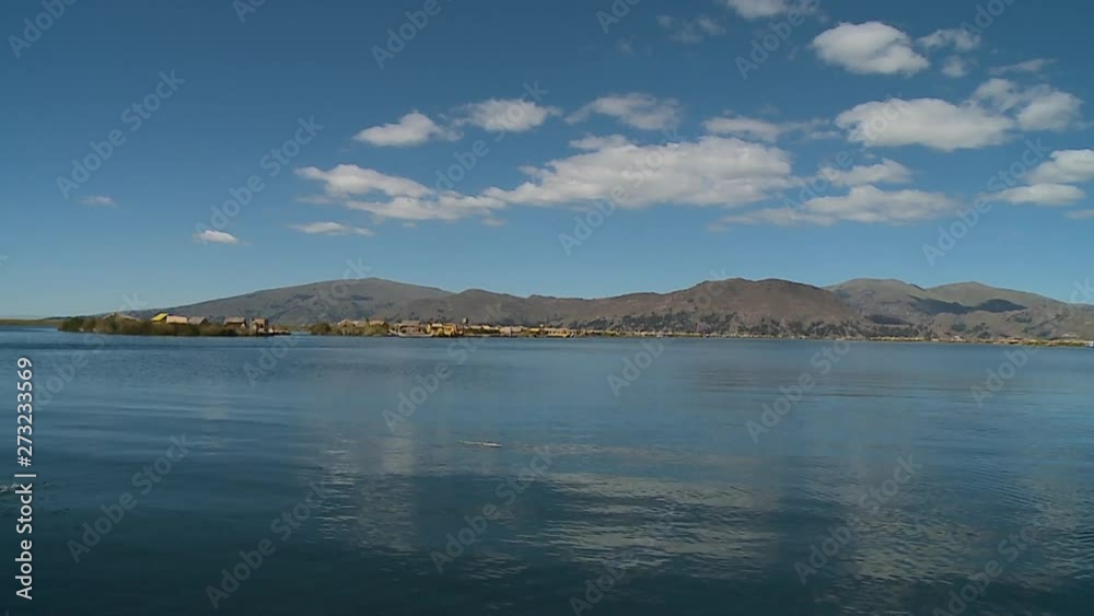 Wide low angle still shot of Uros floating reed villages on the calm water of Lake Titicaca. On the horizon are andean mountain ranges below the blue sky, Lake Titicaca, Peru Wide low angle still shot of Uros floating reed villages on the calm water of Lake Titicaca. On the horizon are andean mountain ranges below the blue sky, Lake Titicaca, Peru
