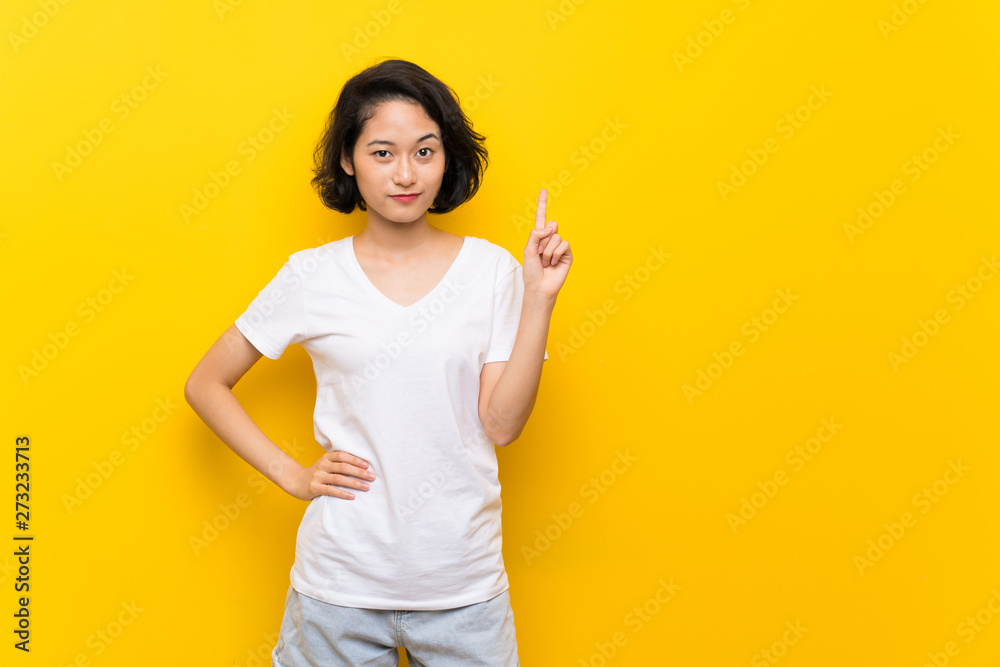 Asian young woman over isolated yellow wall showing and lifting a finger in sign of the best Asian young woman over isolated yellow wall showing and lifting a finger in sign of the best
