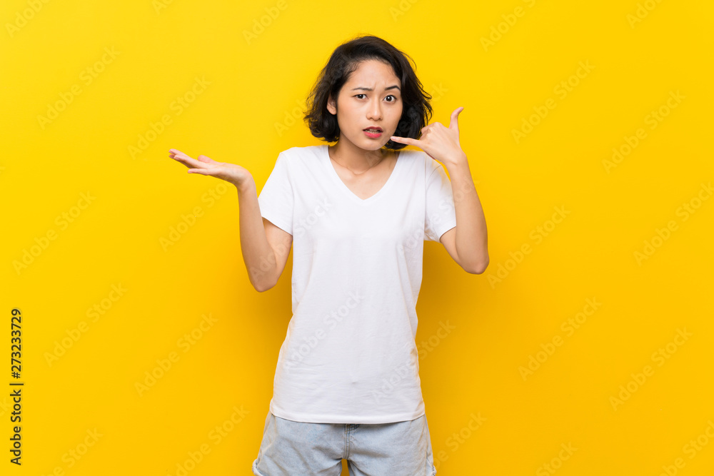 Asian young woman over isolated yellow wall making phone gesture and doubting Asian young woman over isolated yellow wall making phone gesture and doubting