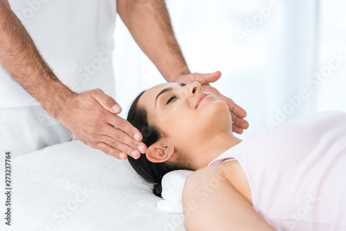 cropped view of healer putting hands near head of happy woman with closed eyes lying on massage table