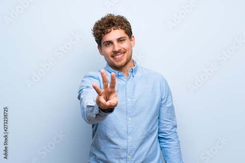 Blonde man over blue wall happy and counting three with fingers