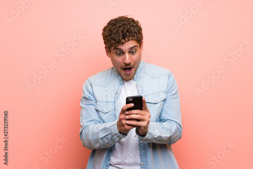 Blonde man over pink wall surprised with a mobile