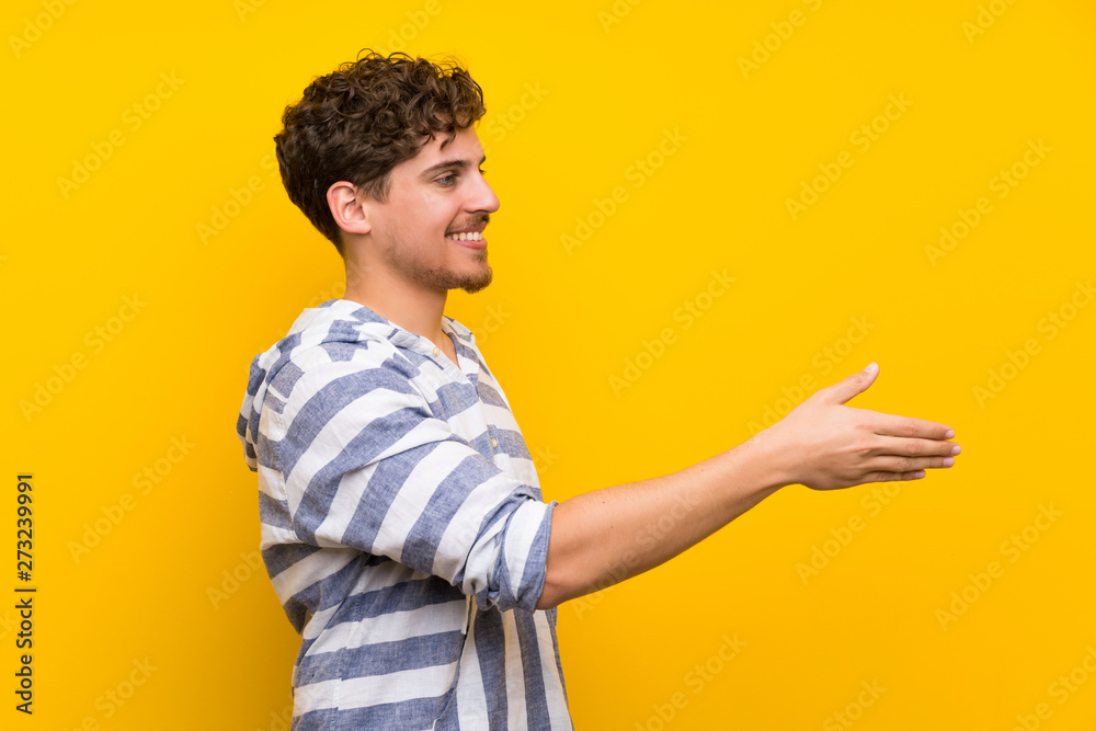 Blonde man over yellow wall handshaking after good deal Blonde man over yellow wall handshaking after good deal