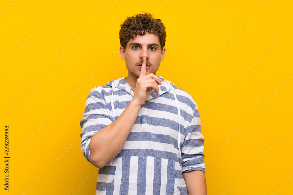 Blonde man over yellow wall showing a sign of silence gesture putting finger in mouth Blonde man over yellow wall showing a sign of silence gesture putting finger in mouth