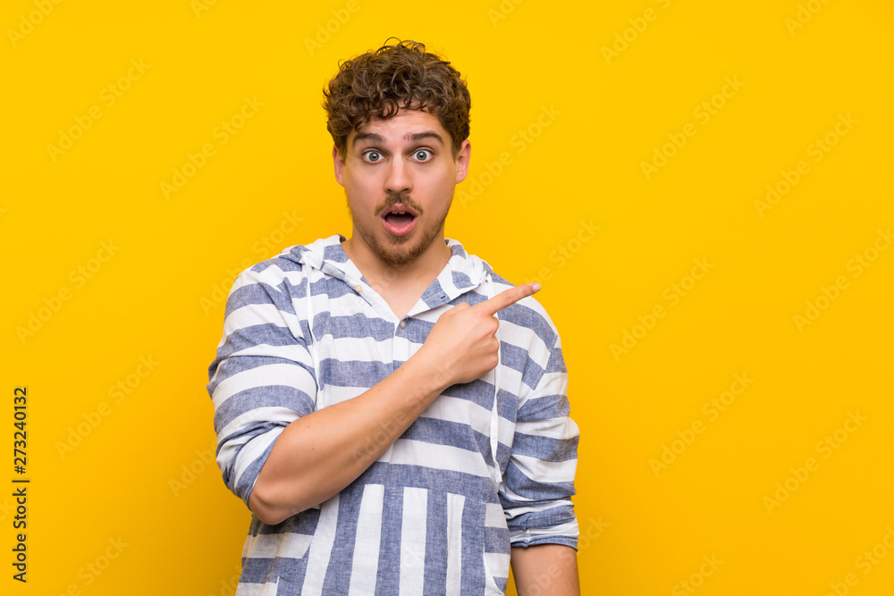 Blonde man over yellow wall surprised and pointing side Blonde man over yellow wall surprised and pointing side