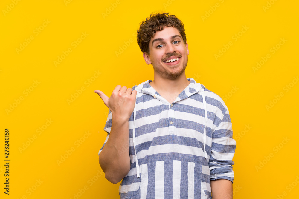 Blonde man over yellow wall pointing to the side to present a product Blonde man over yellow wall pointing to the side to present a product