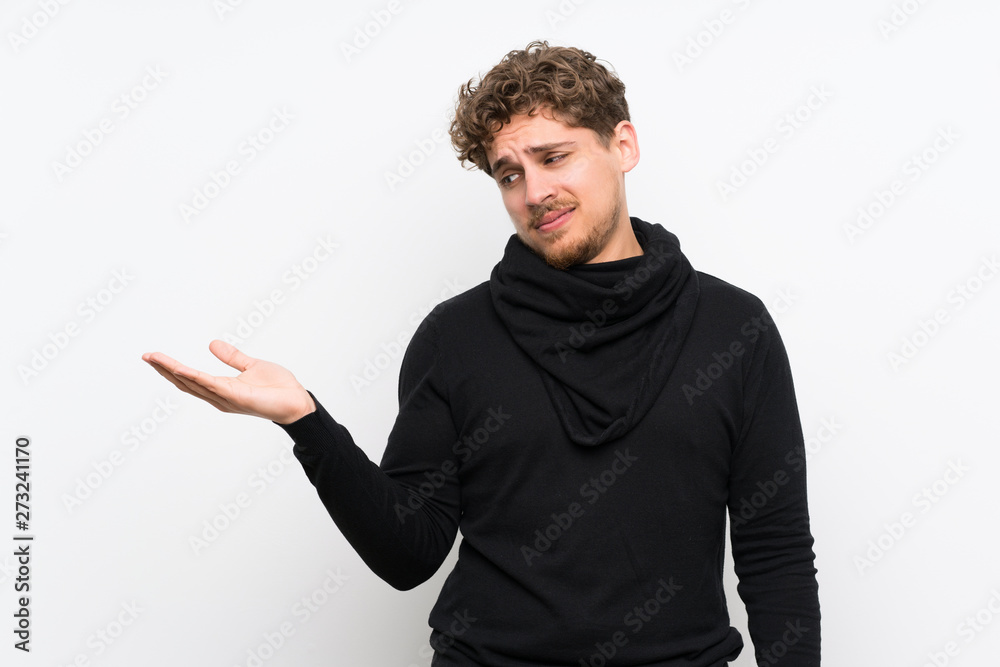 Blonde man over isolated white wall holding copyspace with doubts Blonde man over isolated white wall holding copyspace with doubts
