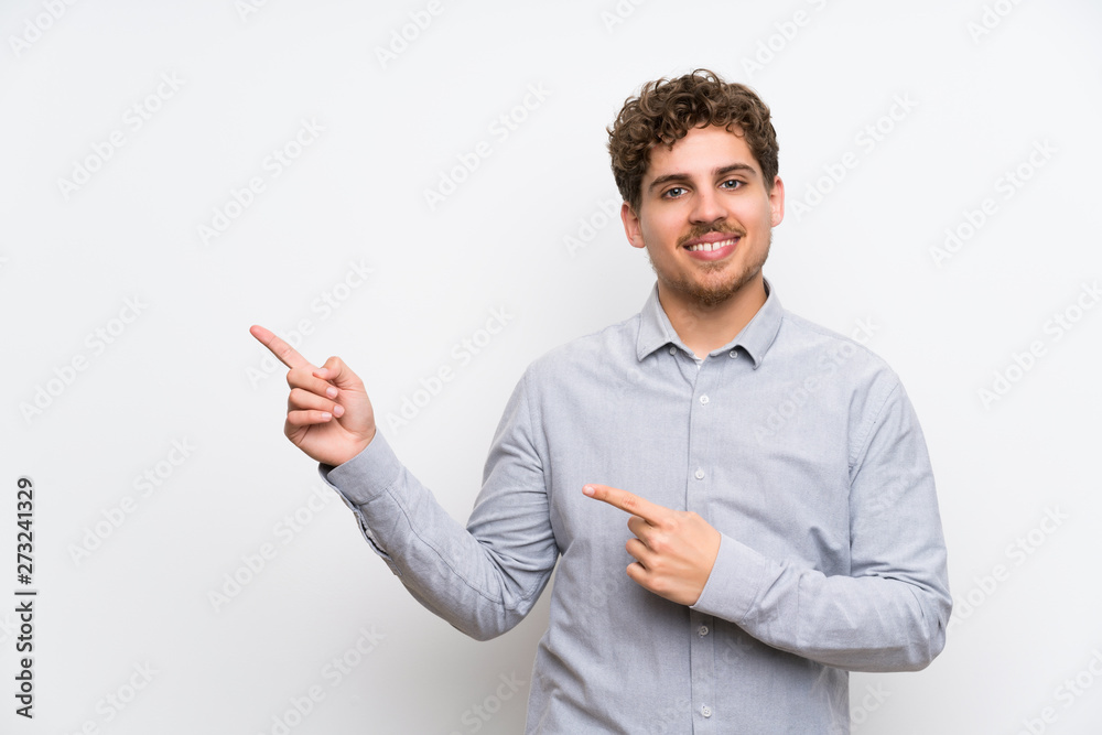 Blonde man over isolated white wall pointing finger to the side Blonde man over isolated white wall pointing finger to the side