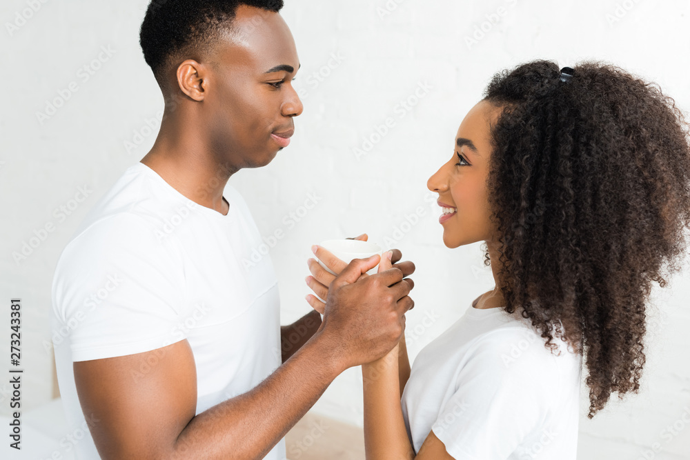 side view of cheerful african american couple looking at each other, holding cup with beverage in hands side view of cheerful african american couple looking at each other, holding cup with beverage in hands