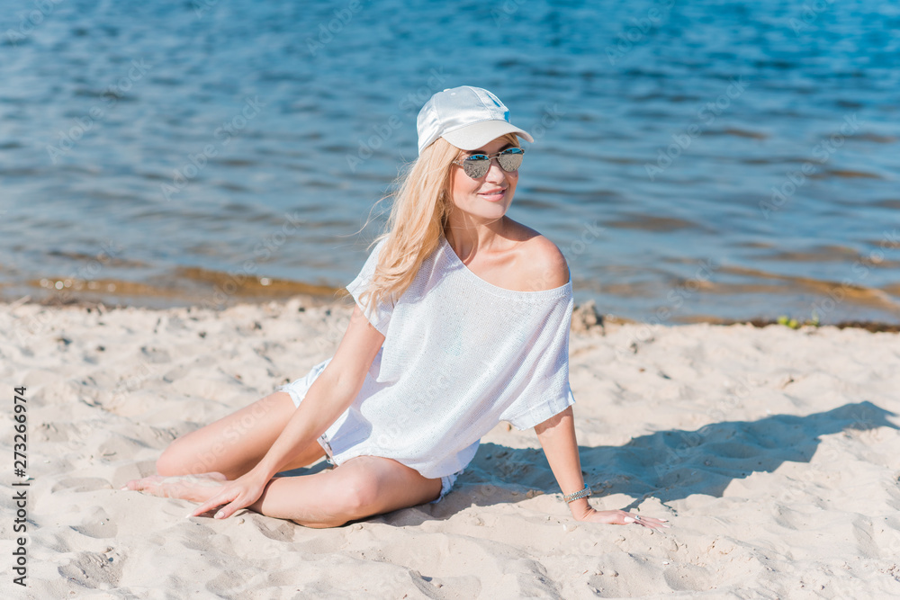 Beautiful nice blonde girl wear white tunic, shorts and baseball cap at the beach, summer fashionable concept 