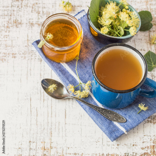 A mug of hot tea, fresh linden flowers and a glass jar with honey on a blue napkin on a wooden table. Prevention and treatment of flu and colds. Selective focus.
