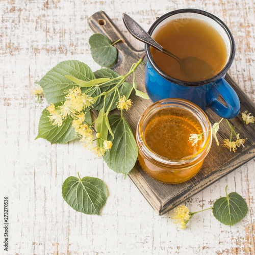 A small glass jar with honey, fresh linden flowers and a mug of hot tea on a square wooden board on a light background. Prevention and treatment of flu and colds. Selective focus.
