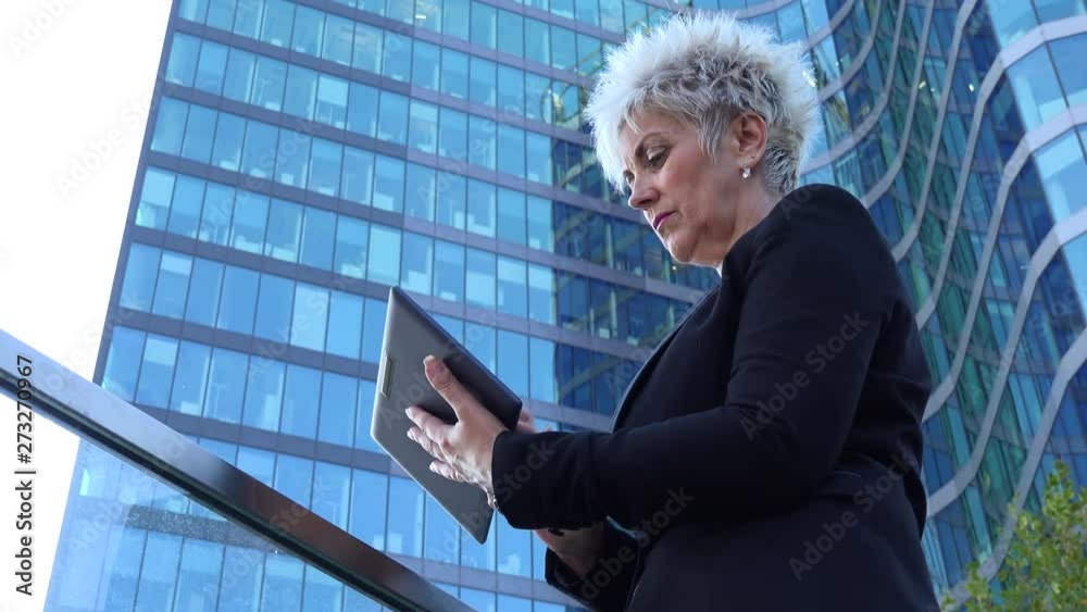A middle-aged businesswoman works on a tablet in an urban area - closeup from below - a windowed office building in the blurry background A middle-aged businesswoman works on a tablet in an urban area - closeup from below - a windowed office building in the blurry background