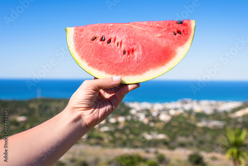 Ripe piece of watermelon in female hands on the background of the sea on a hot summer day. Concept