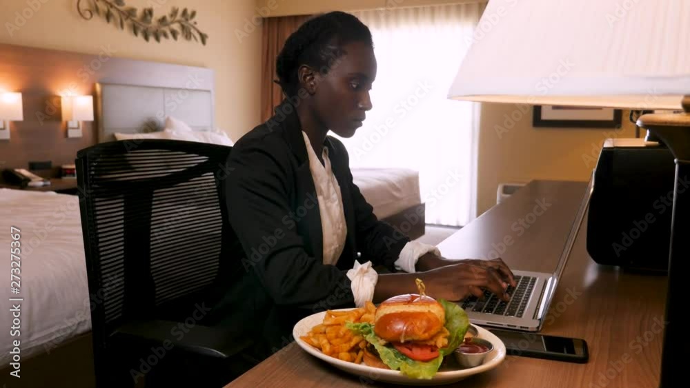 business woman working at hotel desk while eating food. Inside her hotel room, this African decent woman works on her laptop. After typing a bit she picks up a french fry next to her gourmet hamburge business woman working at hotel desk while eating food. Inside her hotel room, this African decent woman works on her laptop. After typing a bit she picks up a french fry next to her gourmet hamburge