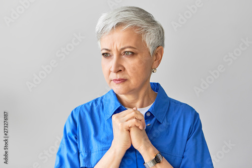 Portrait of serious frowning middle aged mature European woman with gray pixie hair expressing nervousness, holding clasped hands on her chest, being impatient, waiting for results of blood test photo