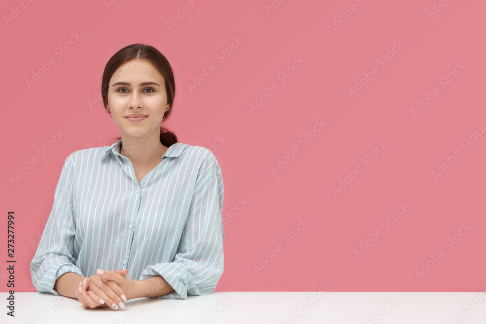 Cute talented young woman in striped shirt sitting at desk with hands clasped during job interview, her look expressing confidence and readiness. Pink background with copy space for advertisement Cute talented young woman in striped shirt sitting at desk with hands clasped during job interview, her look expressing confidence and readiness. Pink background with copy space for advertisement