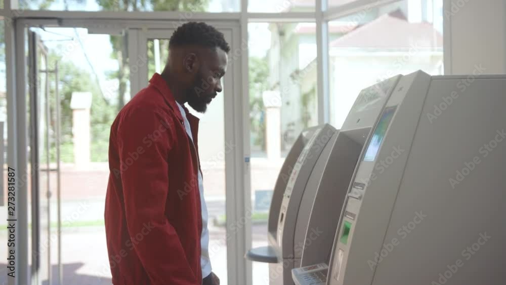 Side view of afro-american attractive man using ATM machine inserting a credit card and entering pin indoors. Side view of afro-american attractive man using ATM machine inserting a credit card and entering pin indoors.