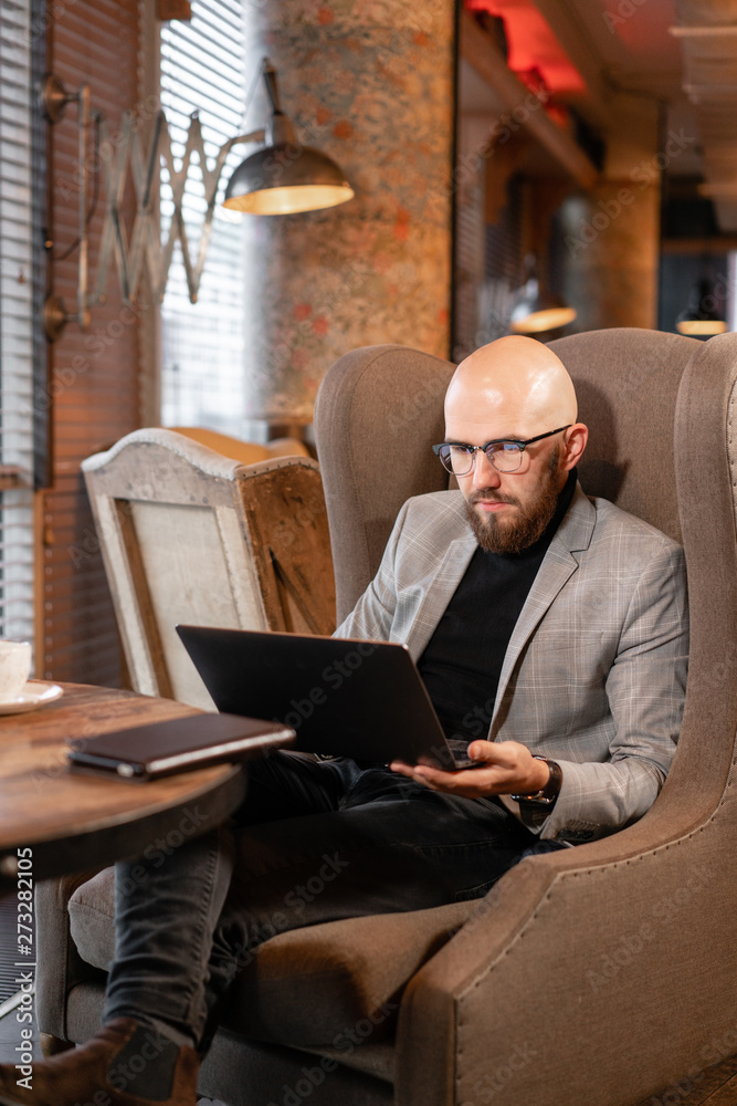 Portrait of young businessman in comfortable cafe. Man with beard in the glasses looking to the monitor laptop. Successful people. Reading text during work on netbook
