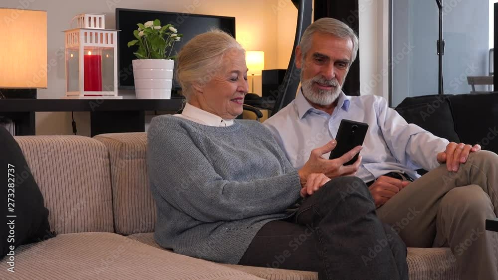 An elderly couple sits on a couch in an apartment, looks at a smartphone and talks with smiles An elderly couple sits on a couch in an apartment, looks at a smartphone and talks with smiles