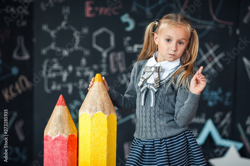Back to school! A little blonde girl in school uniform stands with two very large pencils on the floore against chalkboard with school formulas at school. The kid is studying in the classroom.