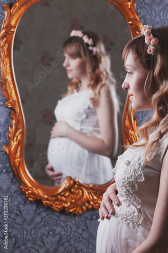 Portrait of a beautiful, young, blond long-haired pregnant mother in a white vintage peignoir, with a floral vink on her head