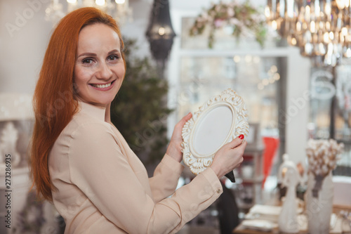 Happy red haired woman smiling to the camera over her shouder, holding photo frame. Attractive mature woman shopping at home decor store. Housewife, interior design concept photo