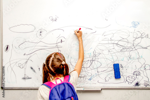 girl with a blue backpack stands with her back and draws a marker on a white school board