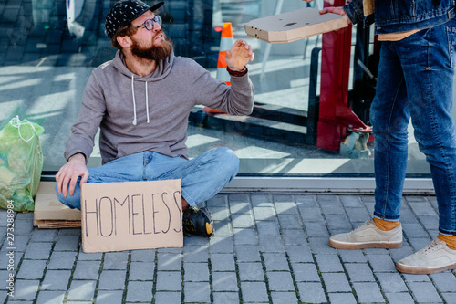 Generous unrecognizable male passerby offering box with pizza for homeless beggar man sitting on the ground with garbage bag near the shopping mall. Concept of human kindness and and sympathy photo