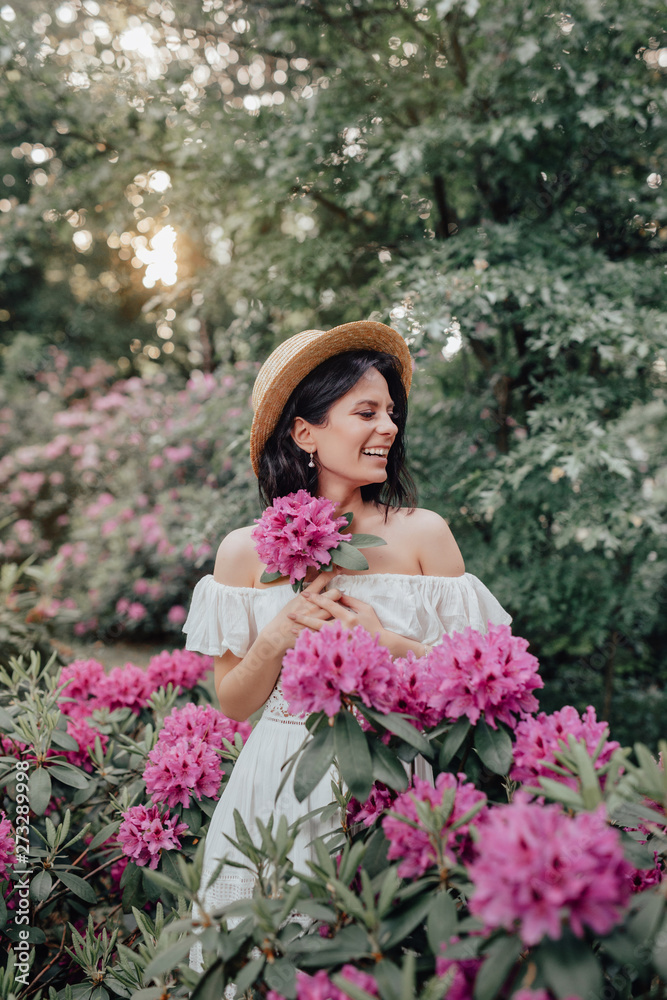 Close up portrait of a beautiful girl in a white vintage dress and straw hat standing near pink flowers