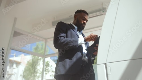 Bottom view of african confident businessman withdrawing a salary from his card. Handsome man in suit inserting a credit card into an ATM machine in a bank. photo
