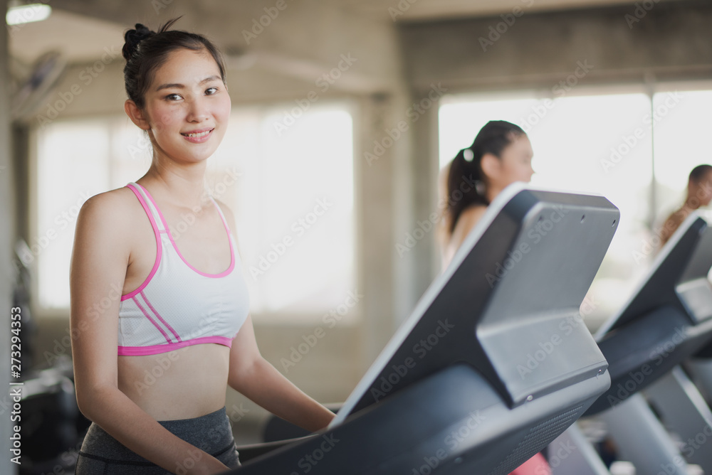 Young Asian woman walking and running on the running machine and looking camera at the gym. Woman workout running at fitness room. People training at gym concept.