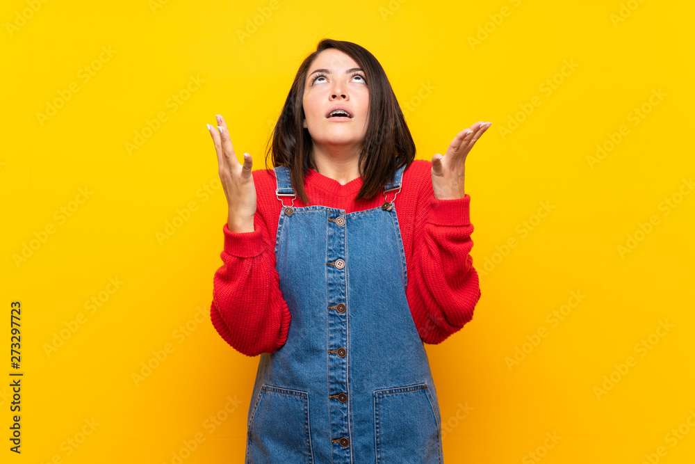 Young Mexican woman with overalls over yellow wall frustrated by a bad situation Young Mexican woman with overalls over yellow wall frustrated by a bad situation
